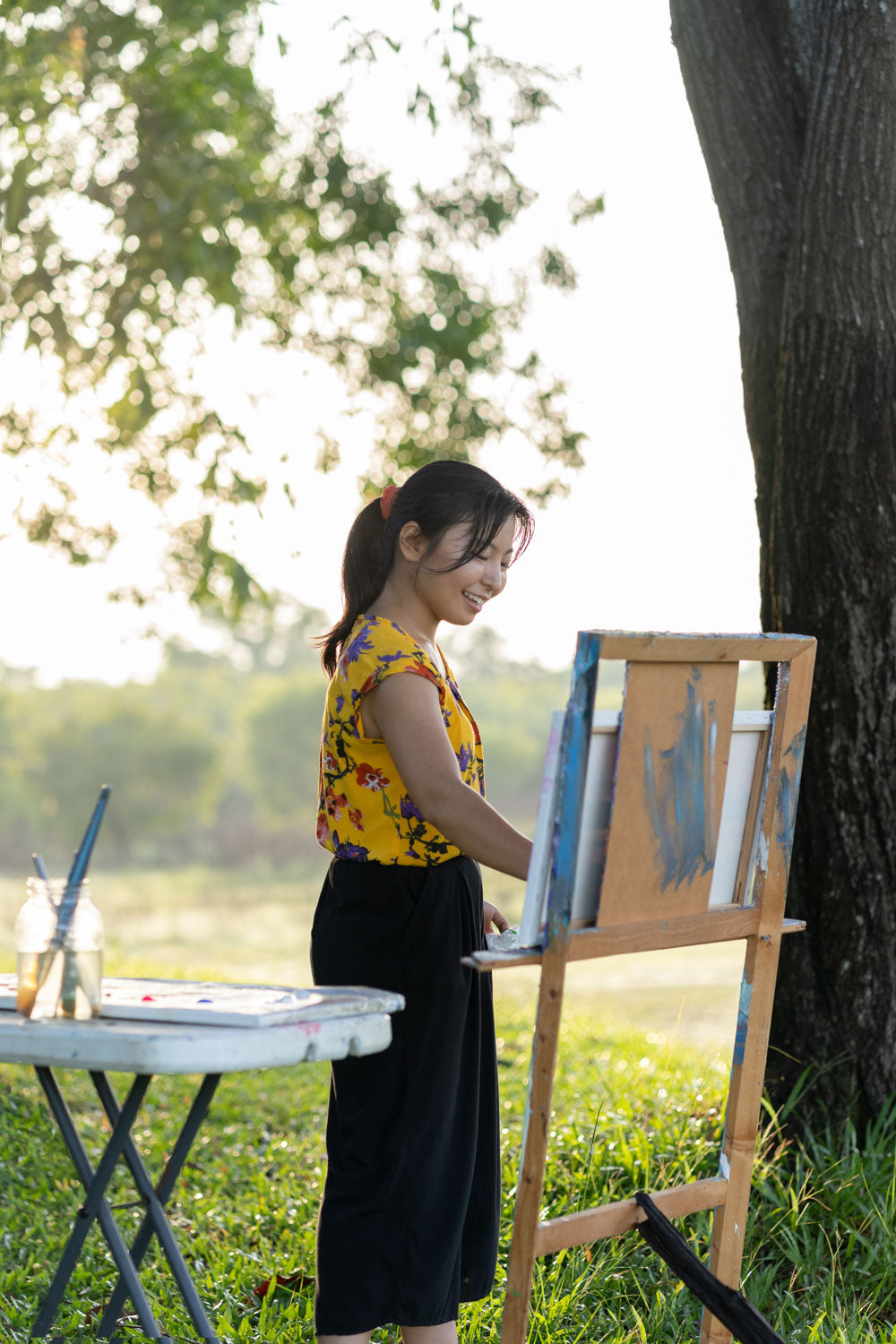 Michiko painting outdoors with a tree and greenery in the background